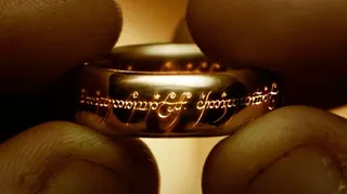 A close-up of Frodo's fingers holding the One Ring, with markings visible on the band.