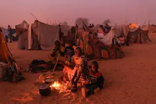 A woman cooks on a small fire outside sitting on the ground. A child sits next to her. Makeshift tents can be seen behind them
