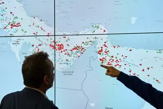 A man looks at a wall map of the strait of Hormuz with dots representing ships, while another points at it