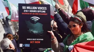 SOPA Images/LightRocket via Getty Images A woman a at a street protest, wrapped in an Iranian flag, holds up a placard. It reads: 90 million voices, 216 + hours offline, silenced by IRGC blackout. Other people holding Iranian flags can be seen in the background.
