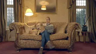 Nicola Baird in jeans, ankle boots and a stripy jumper sits on a patterned sofa with her legs crossed. Her right arm is cuddling her dog. She is sitting in a red-carpeted living room with open heavy curtains at the two windows either side of the sofa.