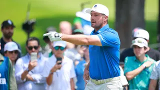 Golfer in a blue polo and white cap points with his gloved hand during a sunny outdoor golf event, with a crowd watching in the blurred background.