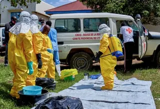 Red Cross workers wearing full-body equipment stand opposite another holding a sprayer and wearing a backpack of disinfectant