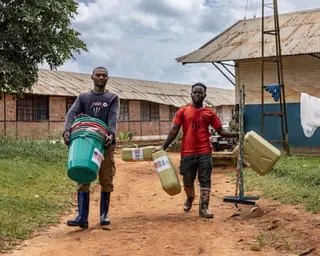 Two men carry plastic buckets and jerry cans as they walk along a path near buildings