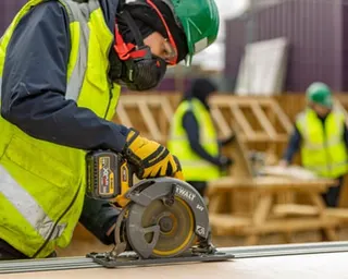 A construction apprentice in high-visibility clothing and a hard hat uses a circular saw