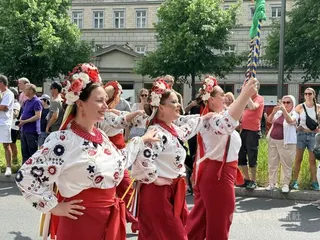 烏克蘭移民頭戴傳統花冠、身穿帶有民族刺繡元素的白色服飾與紅色長裙，在遊行隊伍中隨音樂起舞。花冠與刺繡服飾皆為烏克蘭傳統文化符號。中央社記者林尚縈柏林攝　115年5月25日
