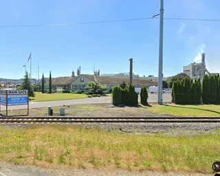 Railroad tracks in foreground with a long industrial building, manicured lawn, and utility pole under blue sky