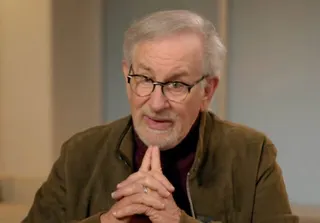 older man with gray beard and glasses in brown jacket looking into camera with two fingers under his chin