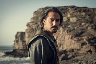 Vilgefortz (Mahesh Jadu) stands in profile on a rocky seashore looking pensively to his right in The Witcher.