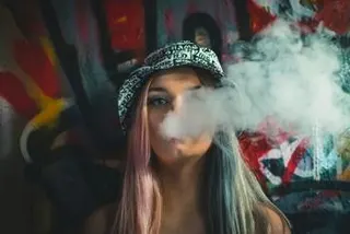 一位留着多彩头发、戴着花纹渔夫帽的年轻女子，在涂鸦墙前吐出一大团烟雾。