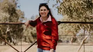 Amy Schultz A woman with long brown hair, wearing a dark red long-sleeved top and jeans, with one arm resting on a large fence with trees in the background
