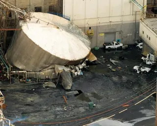 A large white cylindrical tank lies collapsed on its side amid debris and vehicles in an industrial yard