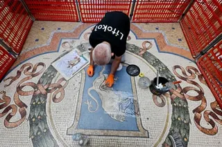 A man in a black T-shirt and orange gloves kneels as he works on the mosaic. The area around it is cordoned off with orange plastic fencing.