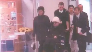 戴著太陽眼鏡和白色針織帽的高英姬坐在輪椅上，由一名男子推著。在法國的一家醫院裡，她周圍有幾個人。