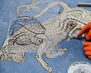 The hands of the restorer, clad in orange gloves, work with tiny tiles on the light brown, cream and pinkish image of the bull, which is laid on a blue background.