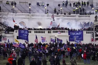 A large crowd gathers in front of the Capitol with smoke visible and Trump 2020 banners displayed