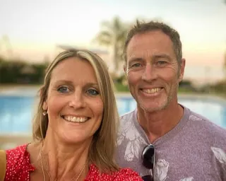 Lindsay and Craig Foreman pose together by a swimming pool with palm trees in the background