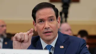 Secretary of State Marco Rubio — wearing a navy jacket, a white collared shirt, a blue tie and an American flag pin — points as he testifies to a House committee.