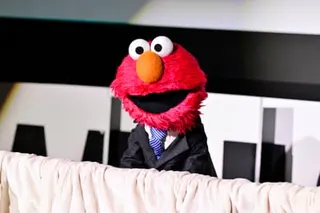 a fuzzy red individual with an orange nose and eyes with no eyebrows speaks on stage while wearing a suit