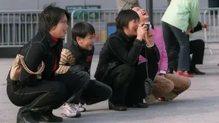 一群人在公共空間深蹲休息，雙腳貼地、膝蓋完全彎曲，其中一人手持相機拍照，背景還有其他站立的人