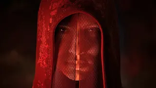 A woman veiled in red gauze and hidden by an ornately stitched red hood stares at the camera. There is darkness all around.