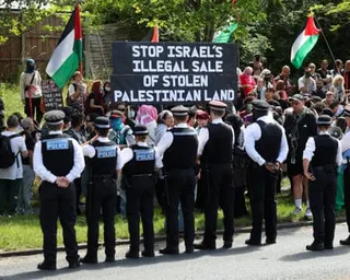 Police officers stand in a line facing protesters holding Palestinian flags and a large sign which reads: Stop Israel's illegal sale of stolen Palestinian land.
