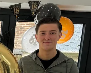 Henry Nowak in an olive green jacket smiles at the camera with 18th birthday balloons and banners behind him