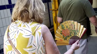 A lady in a white and yellow patterned dress enters a London Underground station with an oriental fan in her hand