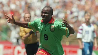 Roger Milla runs as he celebrates scoring against Russia at the 1994 World Cup by spreading his arms out wide with a wide grin on his face. Milla wears a green Cameroon shirt with a red collar and a bold number nine on the chest, with red shorts. Out of focus in the background a referee in a yellow shirt and black shorts and a Russia player in a white shirt and blue shorts can be seen in front of a crowd