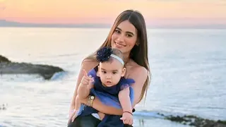 A woman with long dark hair smiles as she holds a toddler in a dress on a beach, the sunset behind her