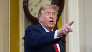 Donald Trump wearing a navy suit and red tie, points his finger to the right side of the frame.
