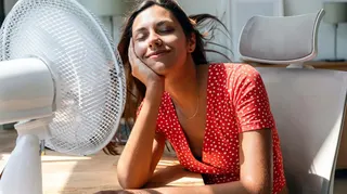 A woman keeps cool by a fan in her home wearing a red dress