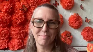 Rosa Thurnher wearing reading glasses with a wall of red flowers and decorative butterflys behind her