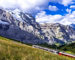 A red passenger train travels along a grassy hillside below dramatic Alps mountain peaks