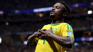 Teboho Mokoena, visible from waist up wearing a yellow South Africa shirt, closes his eyes and leans backwards slightly as he holds his hands across his chest with his fingers interlocked. A packed stadium can be seen out of focus in the background
