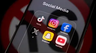 TikTok, Instagram, Facebook, X, Snapchat and YouTube apps displayed on an iPhone screen under a "social media" tab, with an icon used to show under-16s banned reflected across it.
