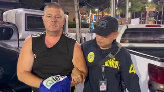Simon Peter Carman, a man with short light brown hair wearing a sleeveless black too, is escorted by a Thai police officer.