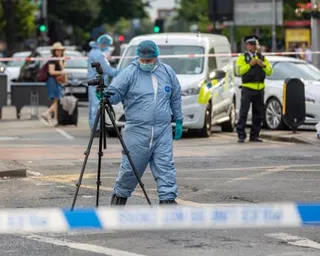 A forensic investigator in a blue protective suit and mask holds a camera on a tripod at a cordoned-off street scene