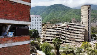 Aerial shot of the side of a damaged red brick building, a partially collapsed building standing opposite. The roof of the collapsed building is partially caved in on the left side