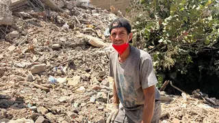 A man in a backwards baseball cap looks into the camera as he stands near a mound of rubble from a collapsed building.