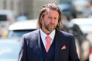 Bearded man with shoulder-length hair in a dark suit and coral tie walks outdoors