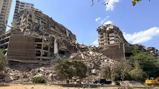Dramatically collapsed buildings behind a dusty roadside lined with trees