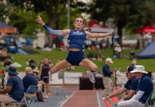 An athlete mid-jump with arms outstretched during a long jump event at an outdoor track meet