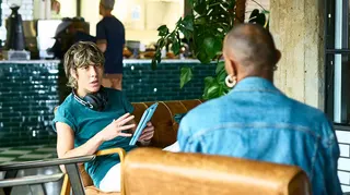 Young man in green T -shirt with headphones around hs neck having a serious discussion with a woman in denim seen from behind. They are in a modern contemporary cafe setting