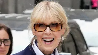 Writer E. Jean Carroll is seen outside looking straight at the camera and smiling. She has blonde hair pulled back and wears large pearl earrings, subglassess and a white shirt with a blue blazer.
