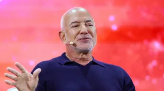 Jeff Bezos wearing a headset microphone and a navy blue polo shirt. He is on stage in front of a bright red background
