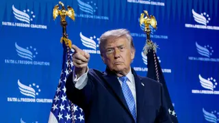 US President Donald Trump pointing a finger, face stern, with American flags behind him.