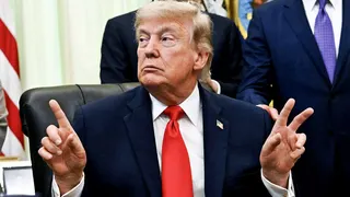 US President Donald Trump pointing fingers in the Oval Office of the White House, with an American flag and suited men behind him.