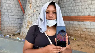 Deilisbeth Herreira stands looking at the camera with a T-shirt on her head and a face mask resting over her chin. She is holding up a photo of her daughters on a phone.