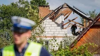 A police officer stands out of focus in the foreground. In the background, people wearing dark clothes and blue latex gloves walk in a hole in a damaged white house, the wooden roof beams exposed.